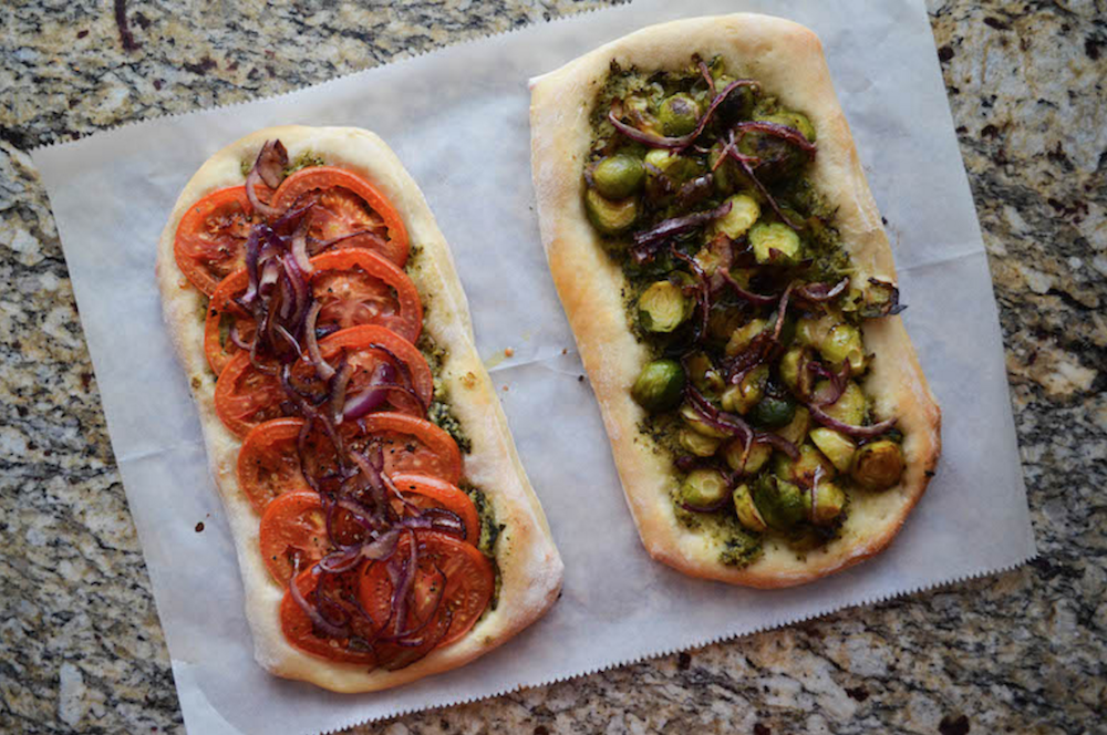 Two flat breads, one topped with sliced tomatoes, red onion, and pesto and the other with caramelized onion, Brussels sprouts, and pesto. 