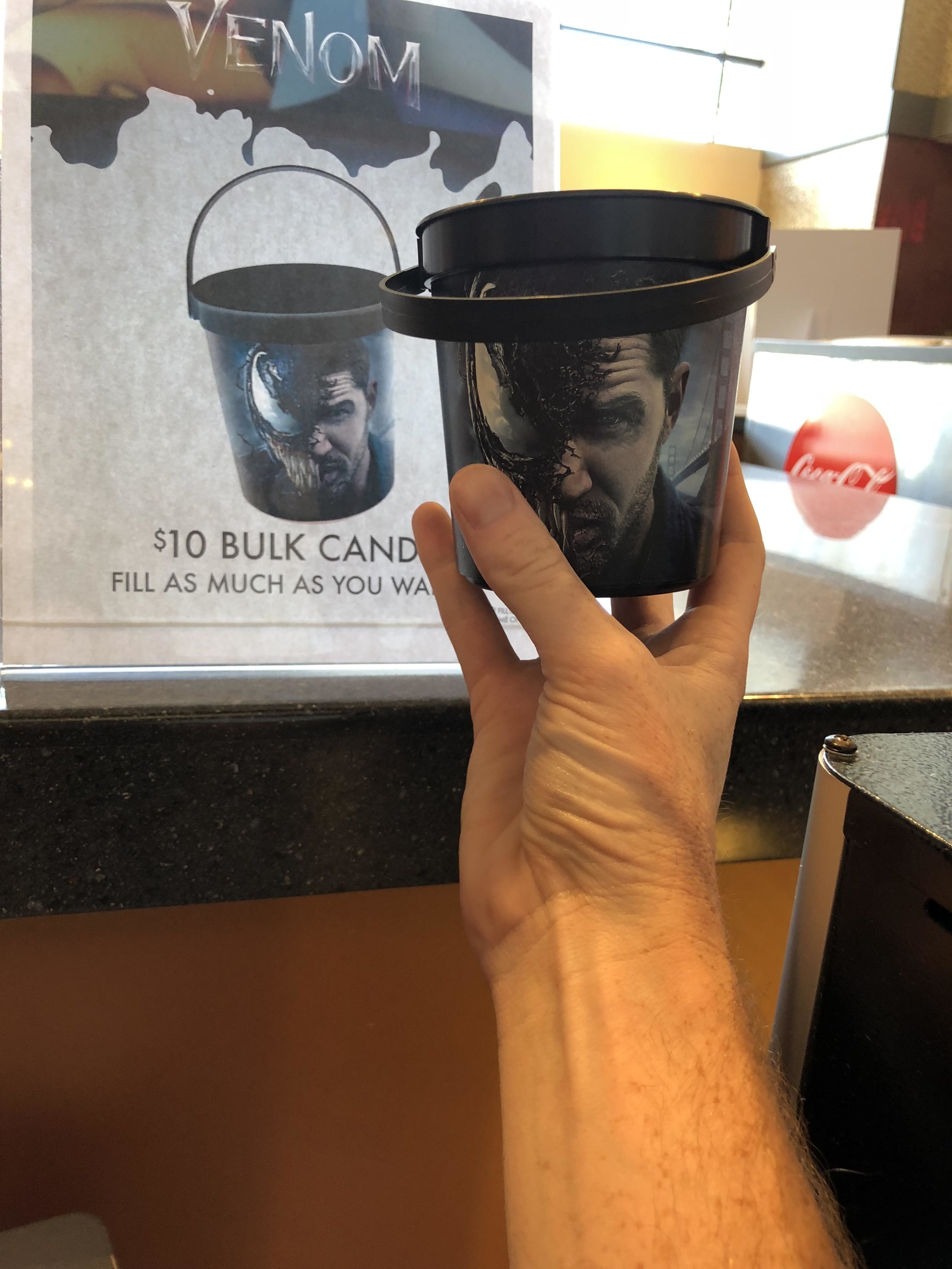 A hand holding up a small bucket in front of an advertisement for the same bucket that reads &quot;$10 bulk candy fill as much as you want&quot;