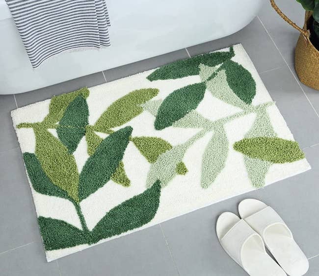 A white bath mat with leaves in three shades of green in front of a bathtub 