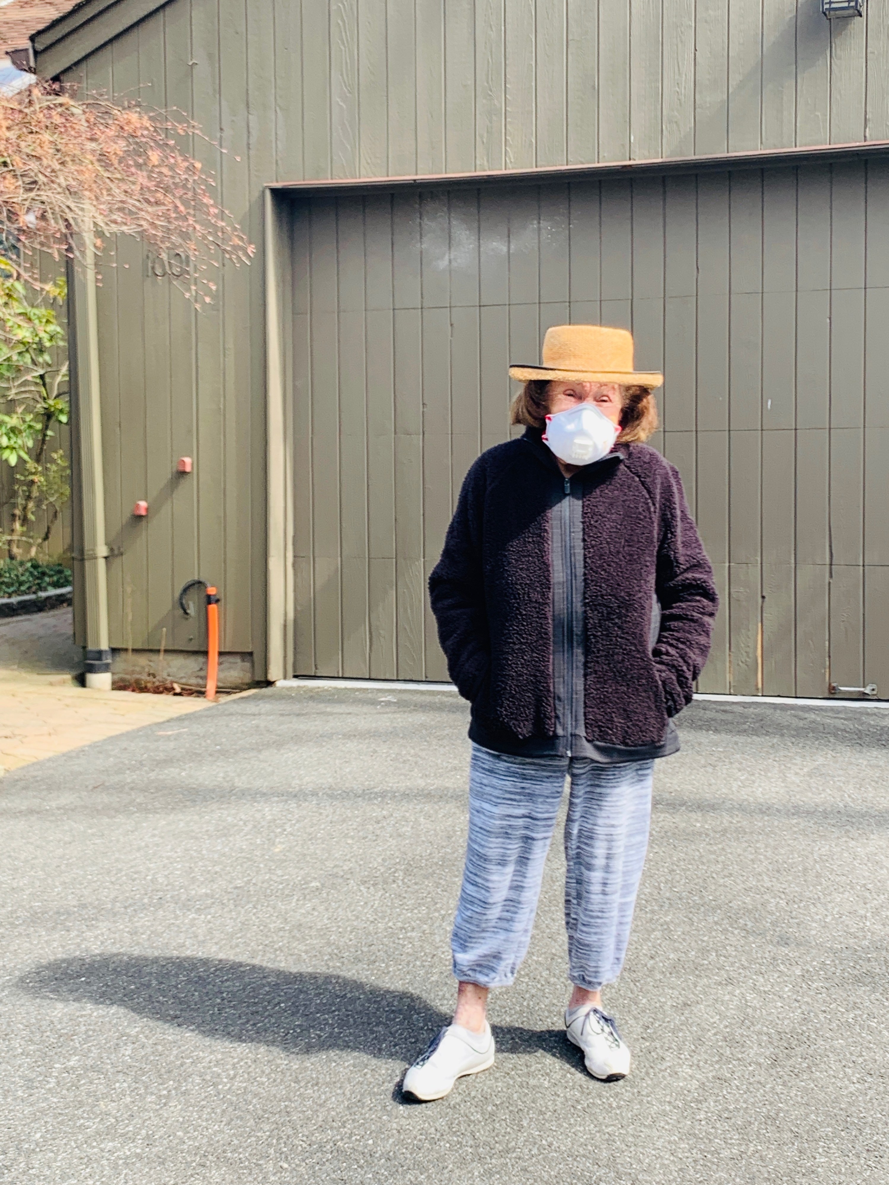 My grandmother standing outside her home in a beach hat and mask.