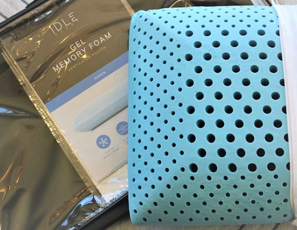 A product shot of the blue memory foam pillow without a pillowcase, sitting beside its packaging. The pillow has cooling gel and is designed with air-circulating holes of different shapes. 
