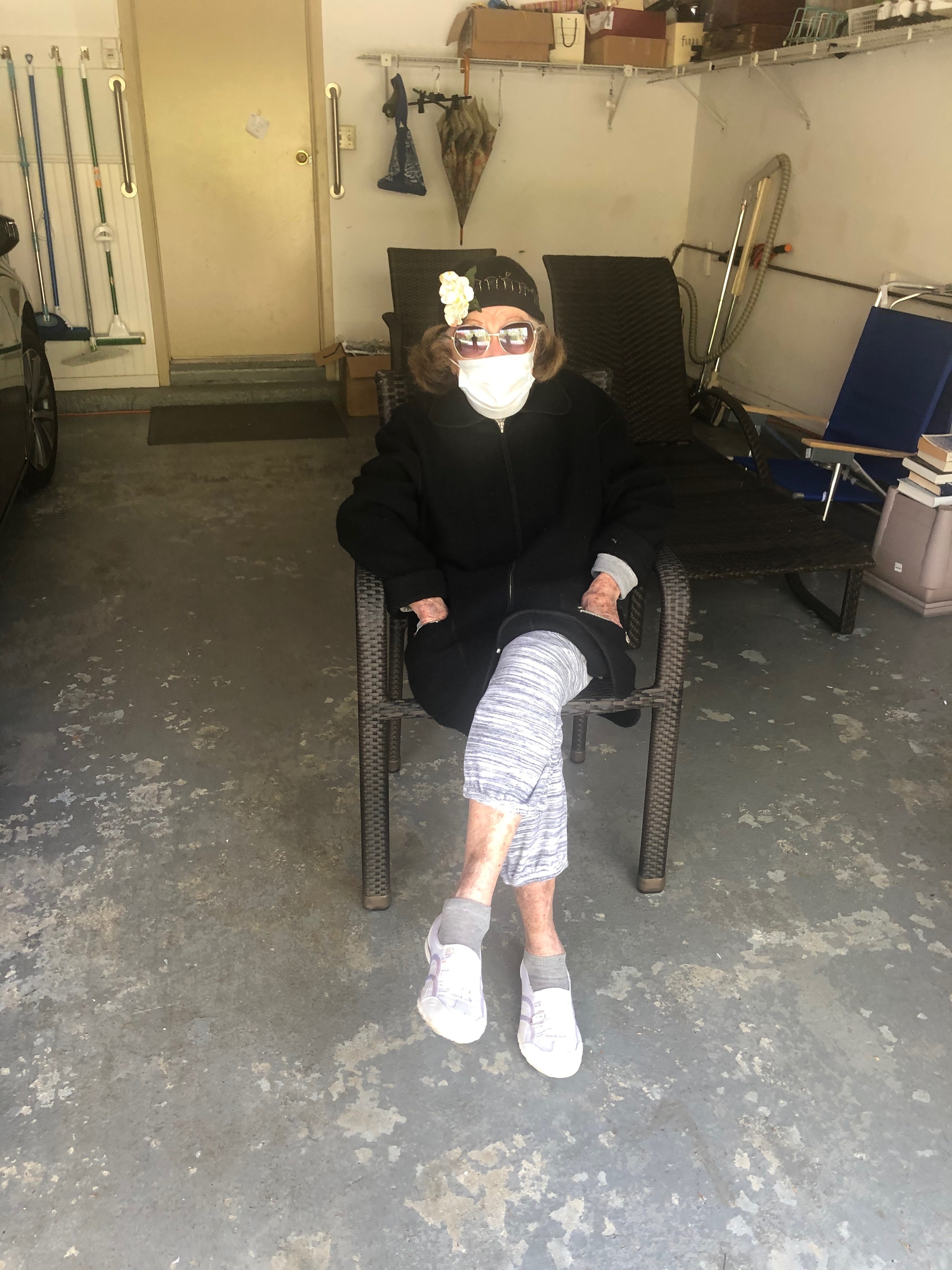 My grandmother sitting in her garage with visitors wearing a mask, a hat, and sunglasses.