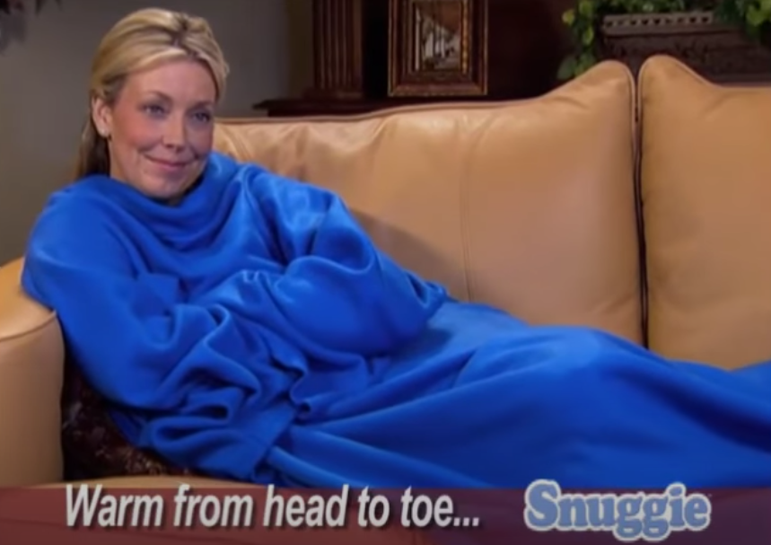 Blonde woman in a blue Snuggie laying on her tan couch watching TV.