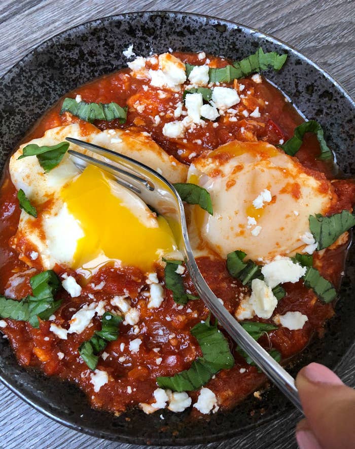 A fork cutting into a runny baked egg stewed in tomato sauce with herbs and feta cheese.