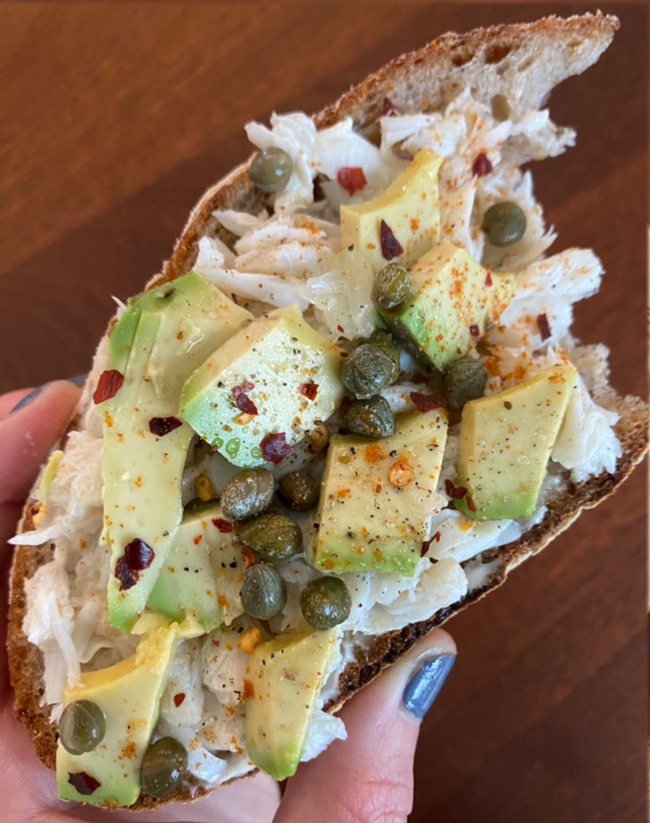 My hands holding a piece of toast topped with crab meat, avocado, capers, and red pepper flakes.