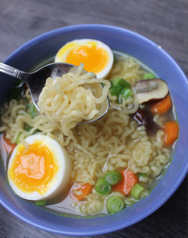 A spoon taking a bite of insant ramen soup with a soft boiled egg, carrots, mushroom, and scallion.