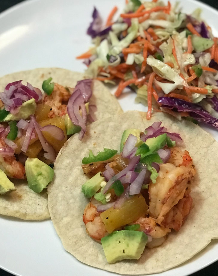 Two tacos filled with shrimp, avocado, slaw, red onion, and cilantro.