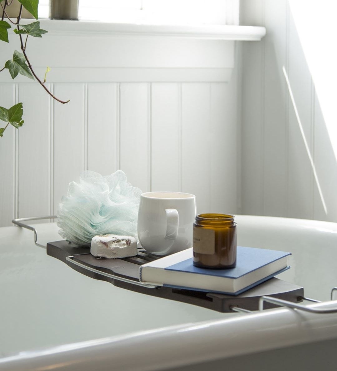 The caddy set on the edge of a tub and topped with a book a candle a loofah and a mug