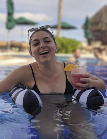 A reviewer sitting in the floatie like a chair with the inflatable arms on both sides of her. She's holdings drink in a pool.