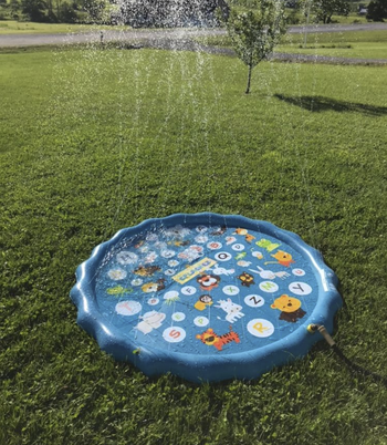 A small inflatable pool with water shooting up from the squirters all around the rim of it. It's sitting in a yard, attached to a hose.
