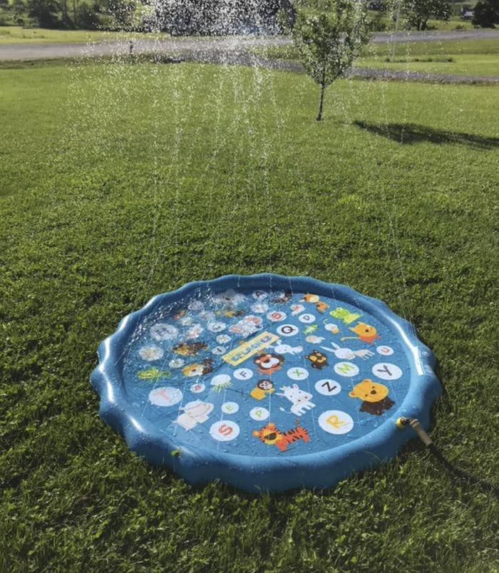 A small inflatable pool with water shooting up from the squirters all around the rim of it. It&#x27;s sitting in a yard, attached to a hose.
