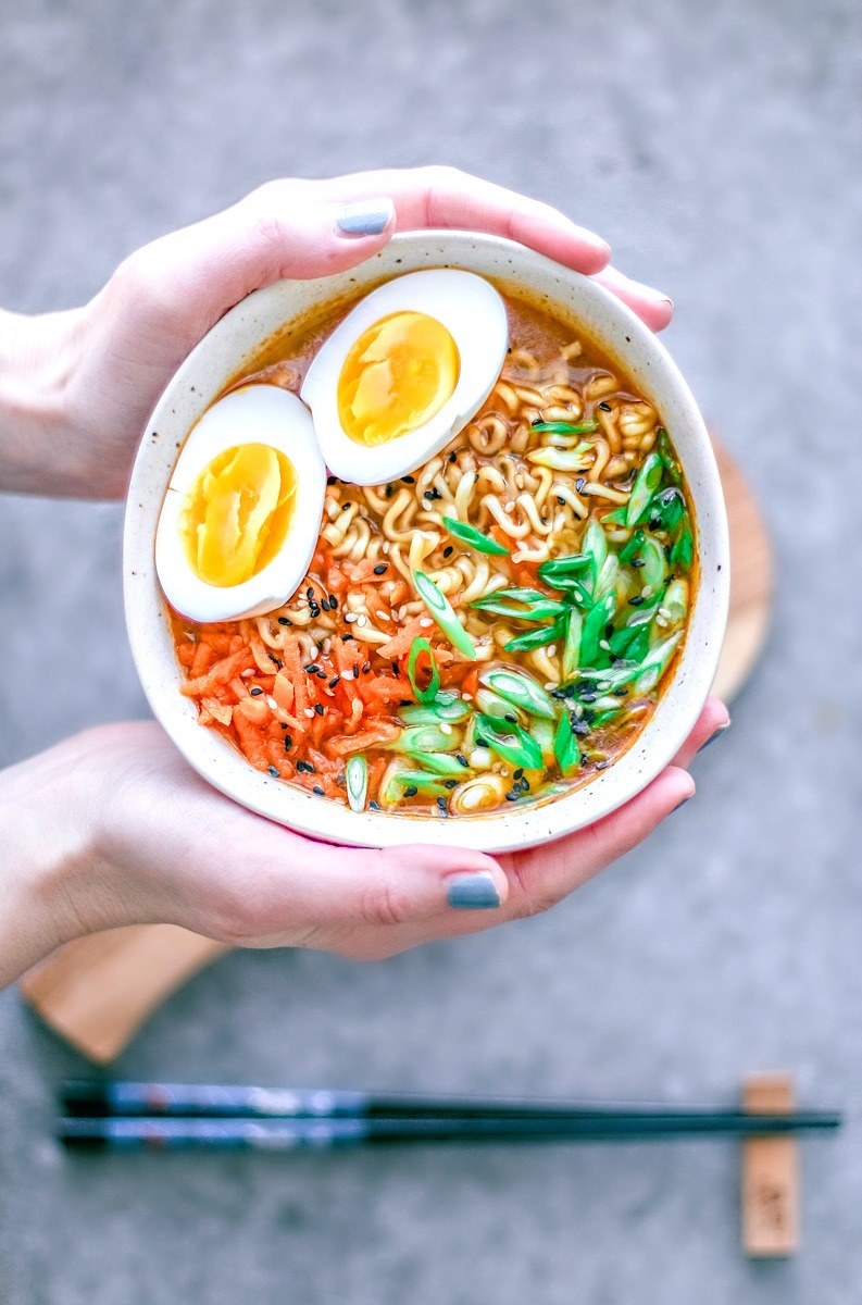 Hands holding a bowl of ramen with chopped scallions, carrots, sesame seeds, and a soft boiled egg.