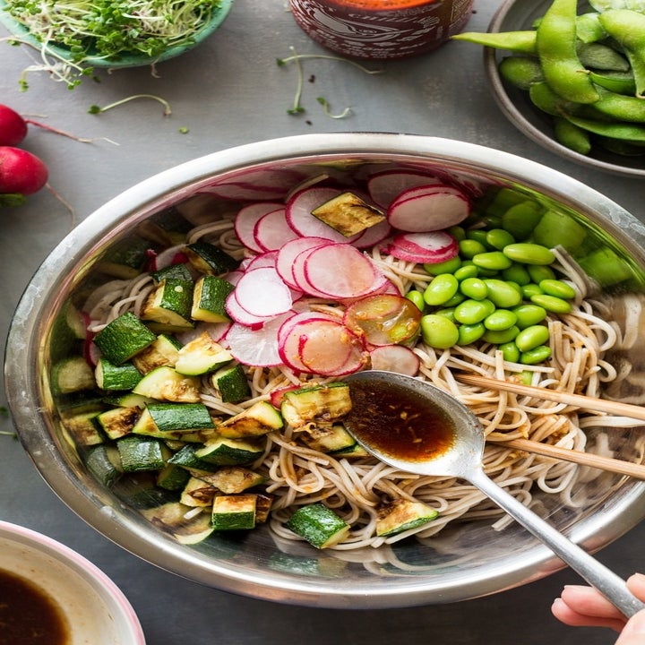A spoon and chopsticks adding soy dressing to a bowl of cold soba noodles with edamame, zucchini, and radishes.