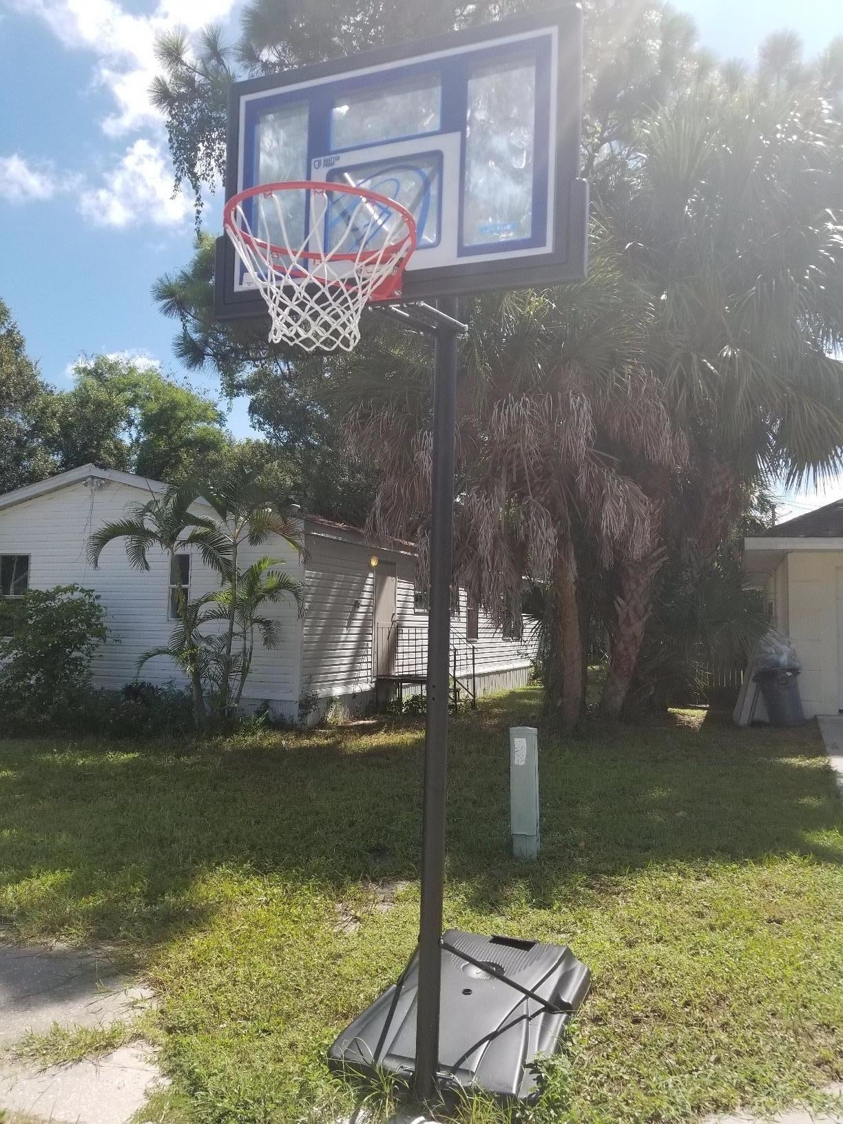 An outdoor basketball hoop with black base and pole, red rim with white netting, and clear blackboard with blue and black accents