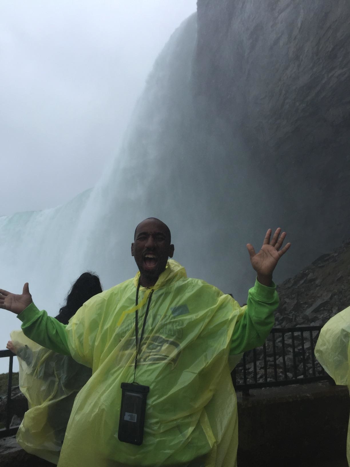 reviewer in poncho excitedly poses in front of niagara falls with phone in pouch around neck 