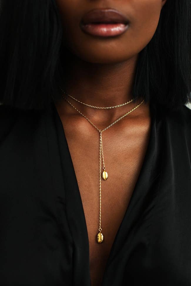 Model wearing the gold necklace. It has two cowrie shells in gold on either end and a long chain that can be worn around the neck or cascading down the chest.
