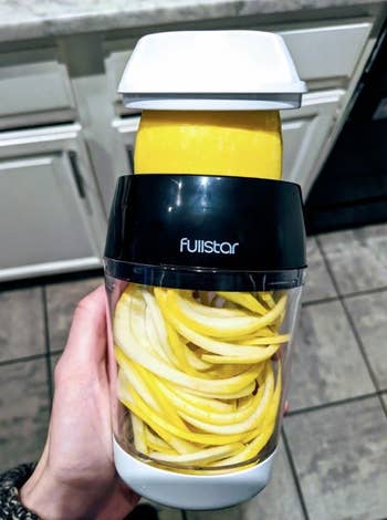 A reviewer holds the spiralizer in their hand, showing a squash mid-spiralized. There is a container attached to the end of the spiralizer, so the zoodles spiral into it