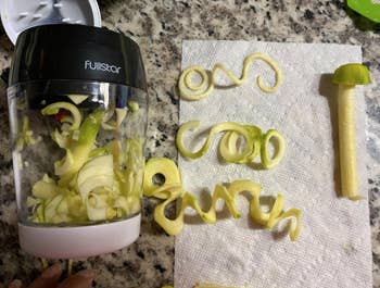 The spiralizer lays on the counter next to a zucchini core, with three zoodle strands spread out to show the three different cuts