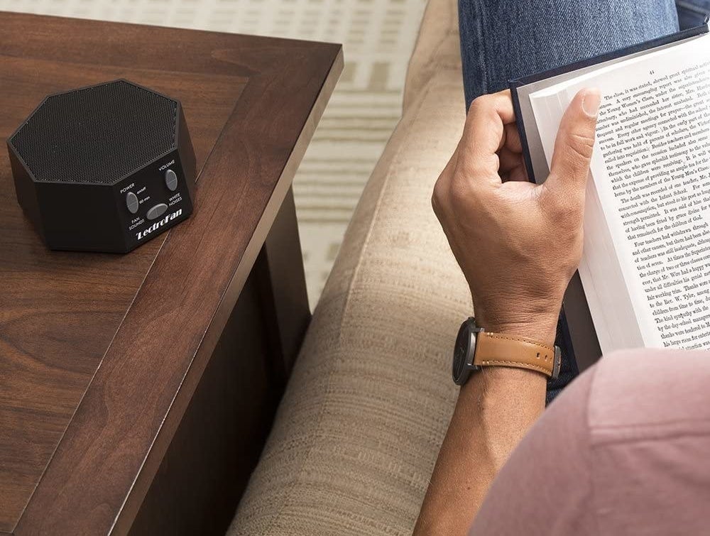 A person sitting on a couch and reading with a small sound machine on the table next to hem