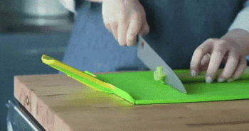 A GIF shows a model chopping vegetables on the cutting board, squeezing the handles, causing the board to fold into a chute, and slide the vegetables on top of a salad
