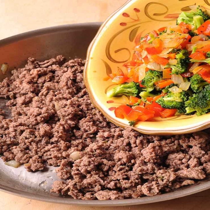 Chopped broccoli, carrots, peppers, and onions being poured into a skillet full of browned ground beef.