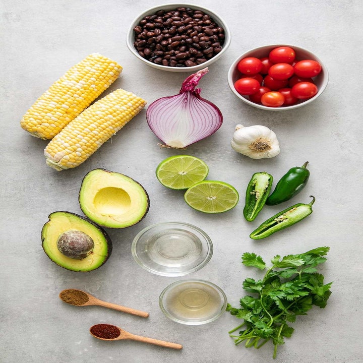 Ingredients on a countertop including corn on the cob, black beans, cherry tomatoes, red onion, lime, jalapeño, and cilantro.