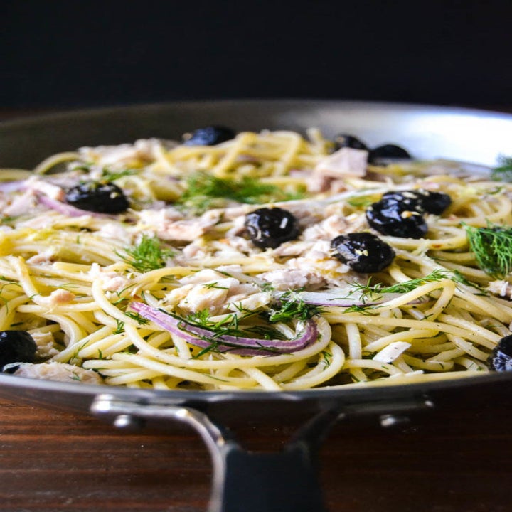 A close-up of spaghetti with canned tuna fish, black olives, and dill in a skillet.