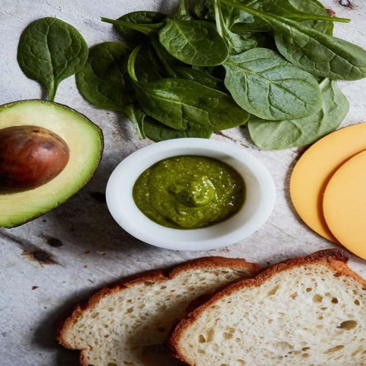 Ingredients on a countertop including two slices of bread, spinach, cheese, avocado, and pesto.