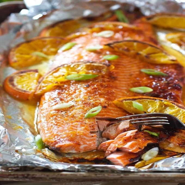 A fork cutting into a flaky piece of marinated salmon topped with orange slices and scallions.