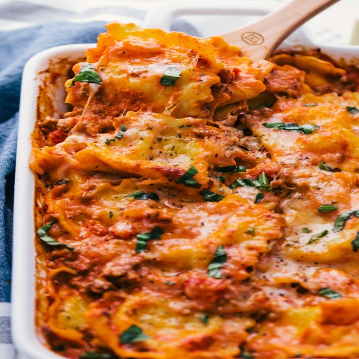 A spoon digging into a baking sheet full of baked ravioli in tomato sauce with fresh chopped basil.