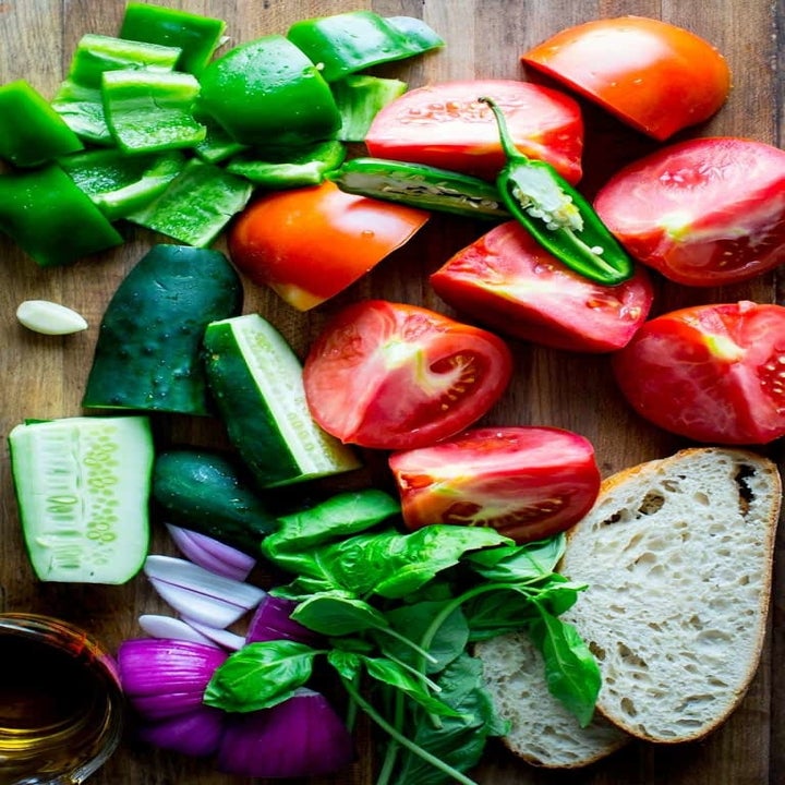 Chopped tomatoes, cucumber, onion, pepper, and bread on a wooden countertop.