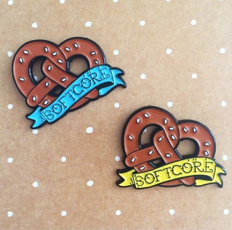 two enamel pins in the shape of a large soft pretzel with "softcore" on a banner across it