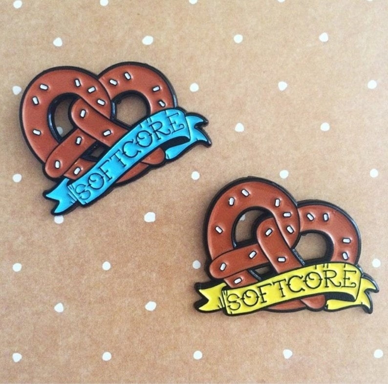 two enamel pins in the shape of a large soft pretzel with &quot;softcore&quot; on a banner across it