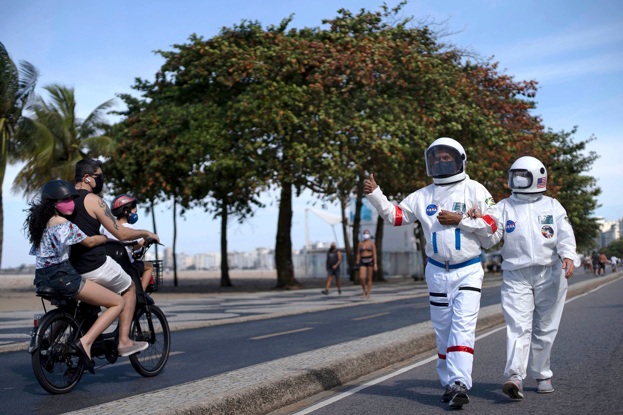 A man and woman wear spacesuits and face masks; the man gives a thumbs up to three people wearing face masks on a bicycle