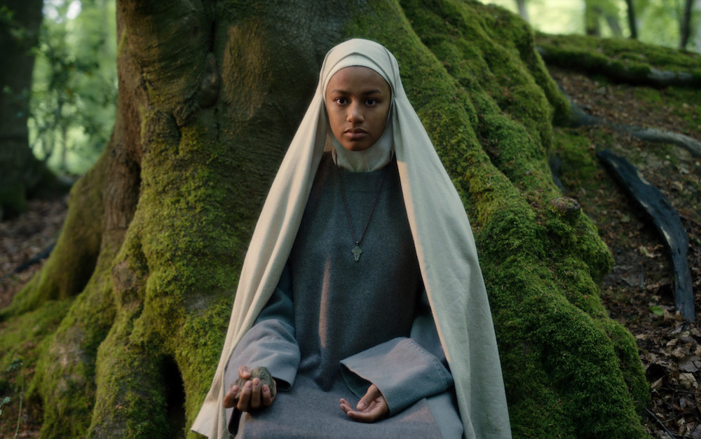 A woman dressed as a nun sits in a forest under a large tree and stares blankly.