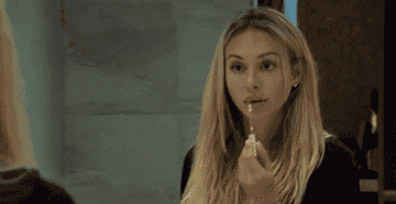 GIF of someone applying lipgloss in a mirror and smacking their lips together 