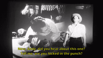 Gif of Michael Stipe dressed as a cowboy walking with a projected film showing Andy Kaufman wrestling behind him and the lyrics now andy did you hear about this one tell me are you locked in the punch on top