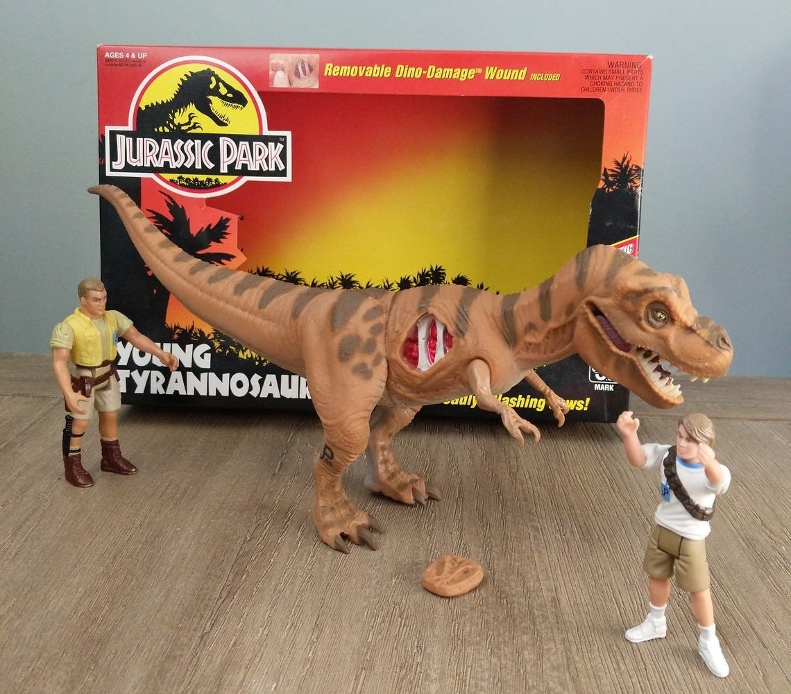 A T-Rex toy with its &quot;dino-damage wound&quot; showing while next to two different Jurassic Park human action figures.