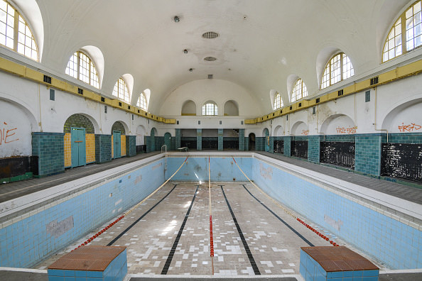 Empty pool in a long gymnasium with high ceilings, broken tiles, and graffiti