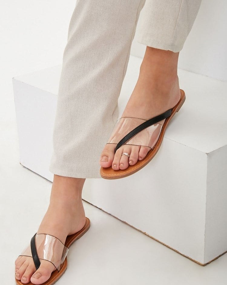 A model wearing the flat sandals, showing the clear cover above the top of their foot and a small black strap that starts at the side of the shoe and crosses over the plastic cover to fit in between the big and middle toe