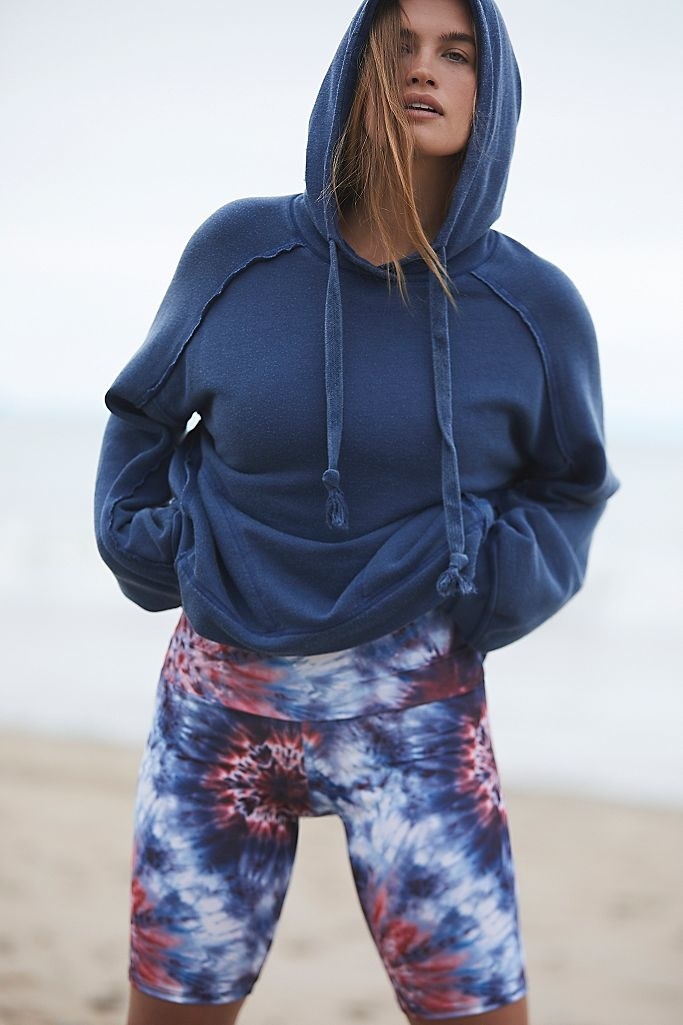 a model wearing the red white and blue tie-dye bike shorts on the beach with a hoodie
