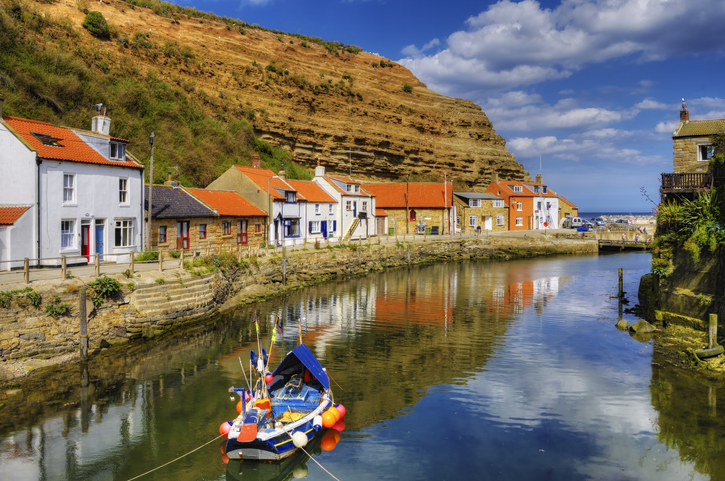 A small fishing boat is on the river in Staithes with the houses either side. You can see the beach and the sea in the distance