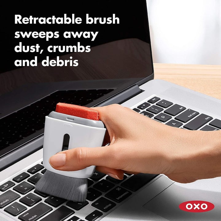 A model's hand using the brush side of the tool to clean their keyboard, with the text "Retractable brush sweeps away dust, crumbs and debris"