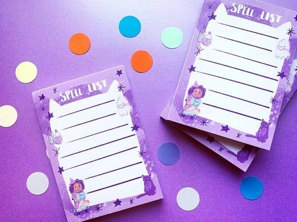 Purple notepads with &quot;spell list&quot; written on the top, lines for notes, and illustrations of a  witch, stars, a ghost, a cat, and potions around the outside