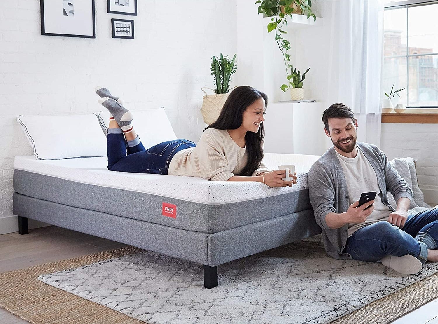 A person lying on a mattress and another person sitting on floor and leaning against the bed frame