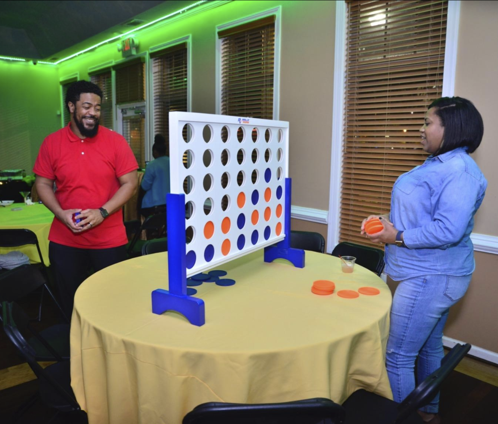 an oversized connect four game on a table which has several holes in it where chips can be inserted via the open top slot. Two people are standing on either side of it playing the game.