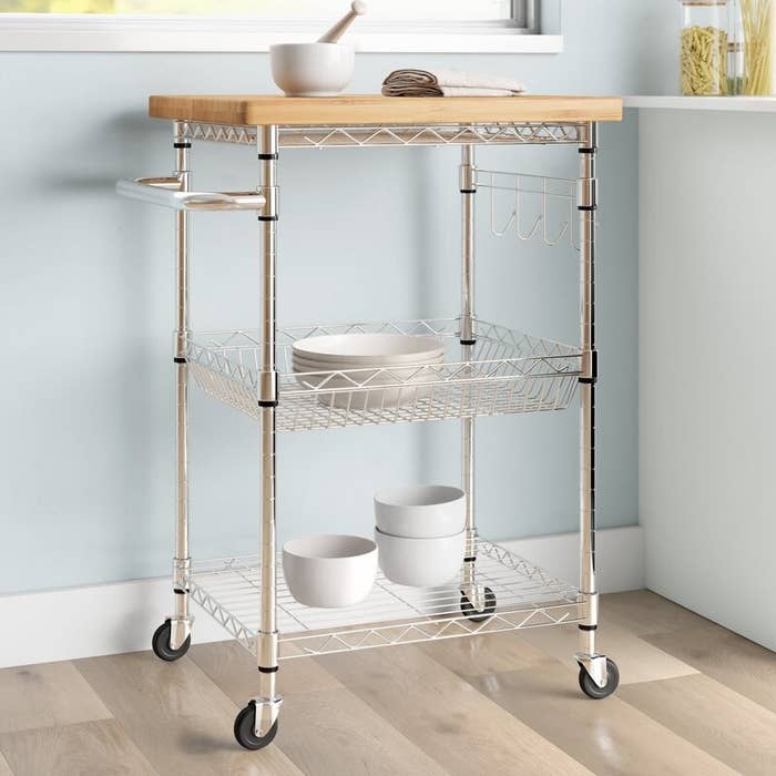 A metal frame kitchen cart on four caster wheels with two shelves and a wooden top