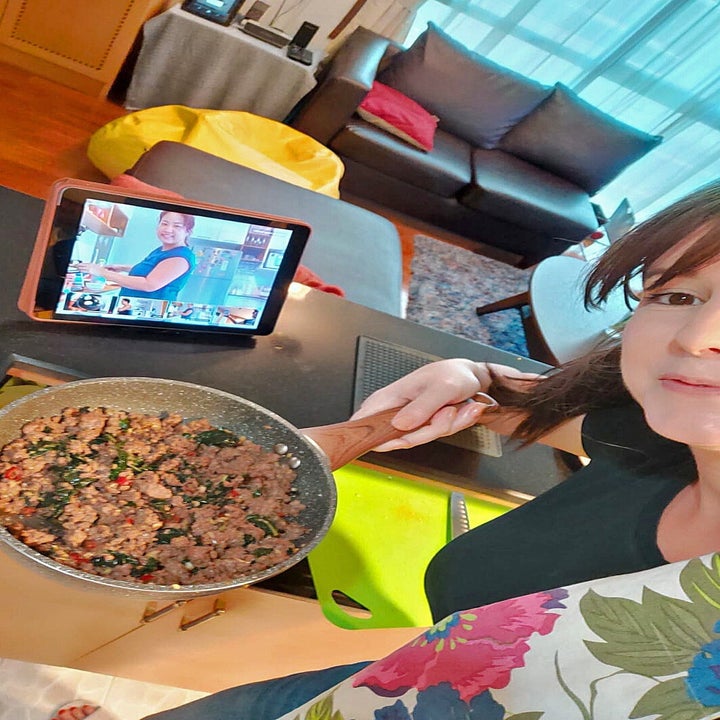 woman taking a selfie in the kitchen while holding up a pan of stir fry. a tablet streaming the cooking class sits on the bench