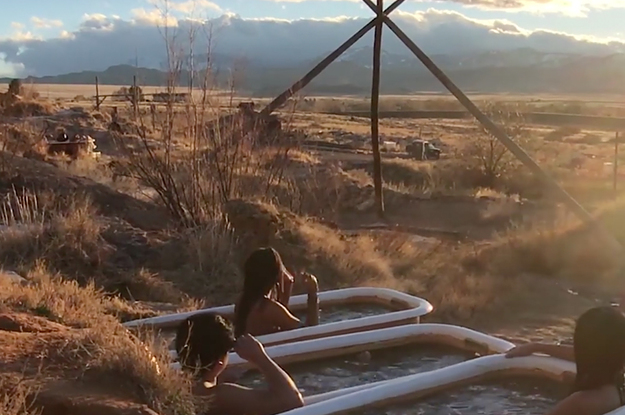 Three people look out over a golden hour desert landscape while soaking in bathtubs embedded into a cliffside.