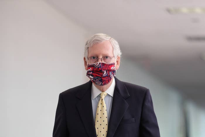 McConnell stands alone in a white hallway wearing a suit and a red-and-blue Washington Nationals-themed mask.